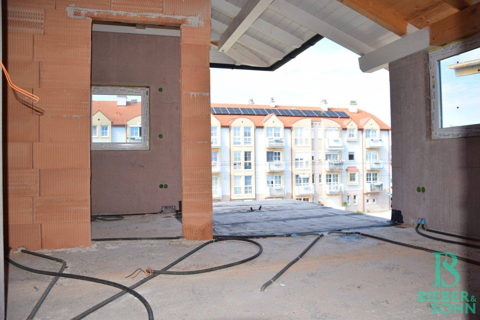Unfertiger Innenraum mit Blick auf ein gegenüberliegendes Gebäude mit Solaranlage und sichtbaren Leitungen.