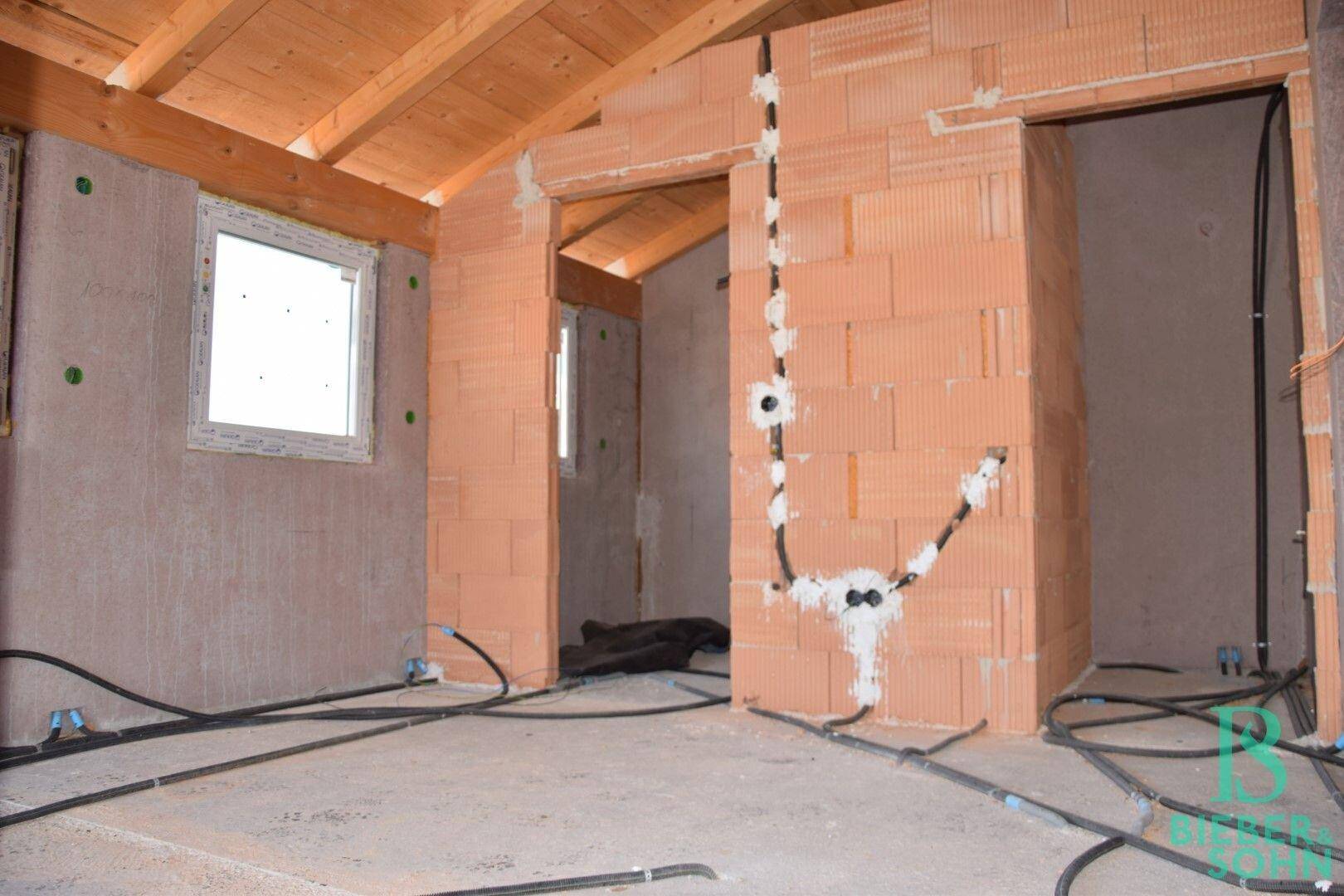 Unfertiger Raum mit Ziegelwänden, Holzdecke und sichtbaren Elektroinstallationen im Rohbau.