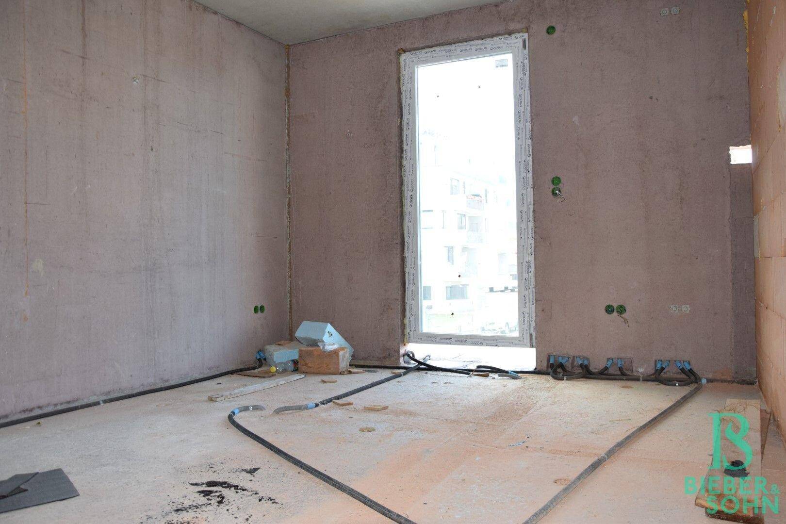 Unfertiger Raum mit großem Fenster und sichtbaren Leitungen am Boden für Fußbodenheizung.