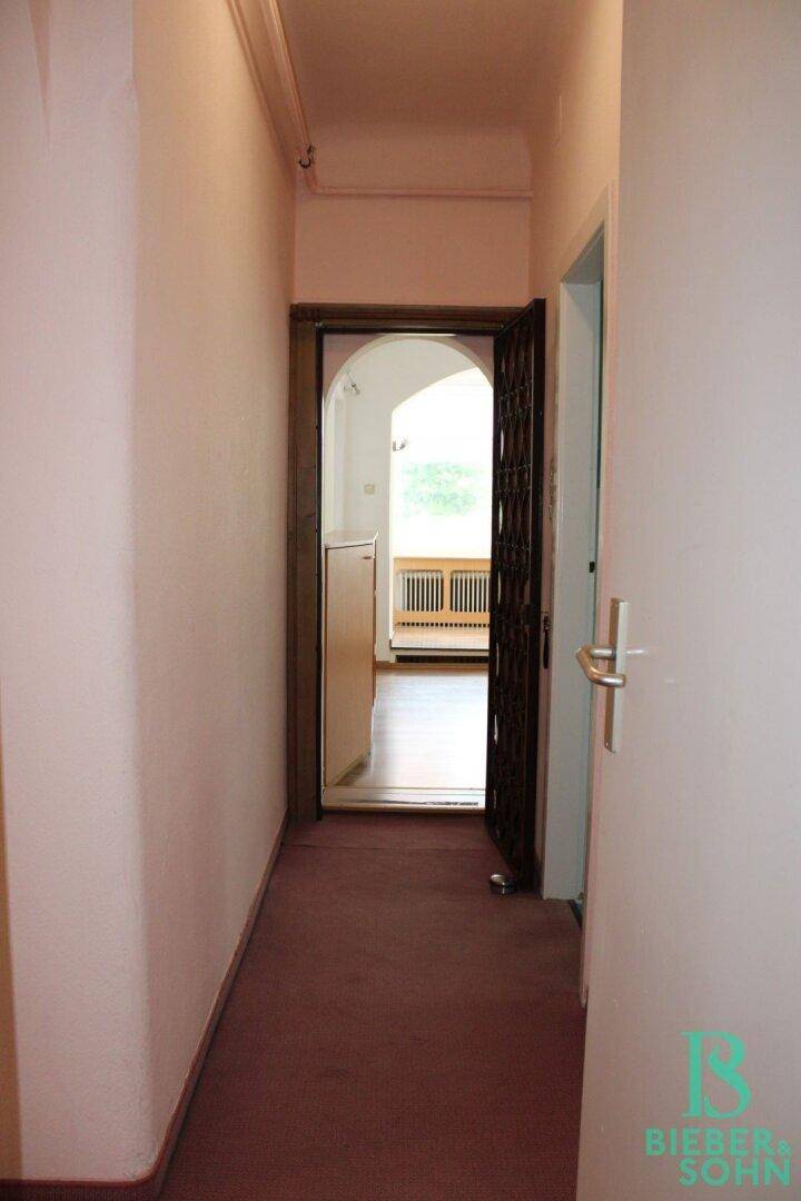 Langer Flur mit Teppichboden und einer offenen Tür, die zu einem hellen Raum führt.