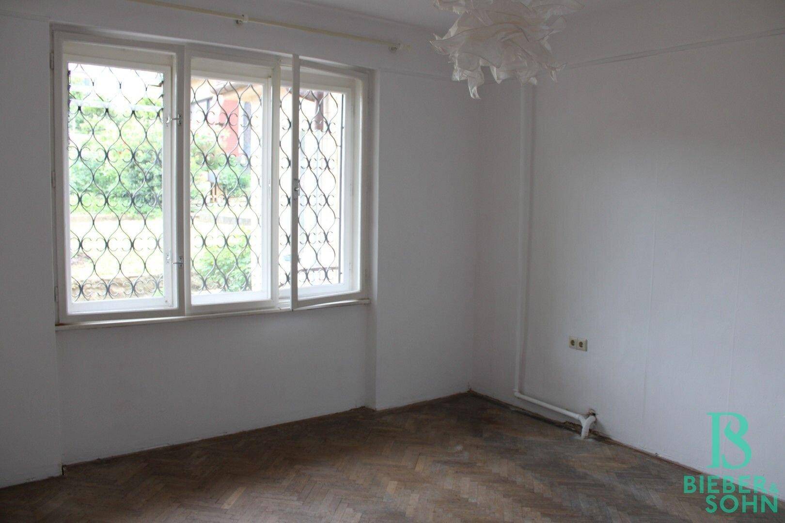 Zimmer mit Fischgrätparkett, weißen Wänden und Fenstern mit dekorativen Gittern.