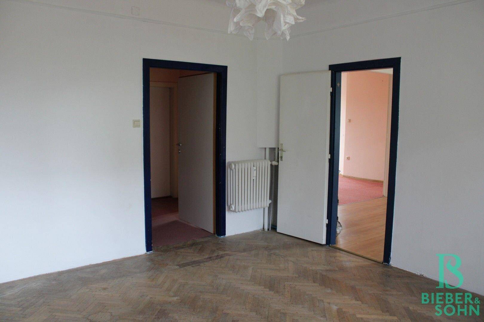 Zimmer mit Fischgrätparkett und zwei Türen, die zu anderen Räumen führen.