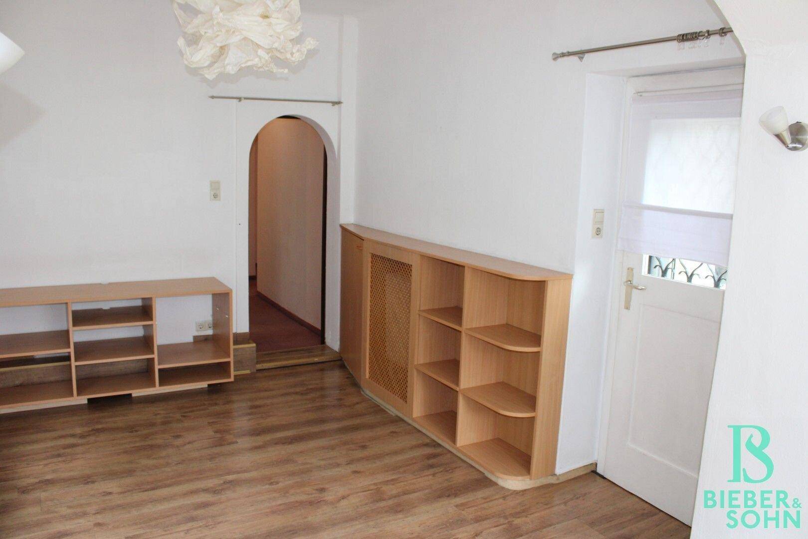 Zimmer mit Holzboden, Einbauregalen und Zugang zu einem weiteren Raum.