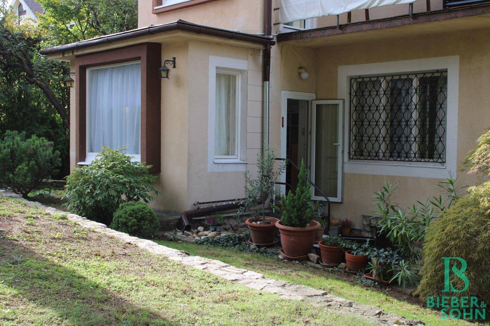 Außenansicht des Gebäudes mit einem kleinen Gartenbereich und Fenstern. Die Fassade ist in einem hellen Beigeton gehalten.