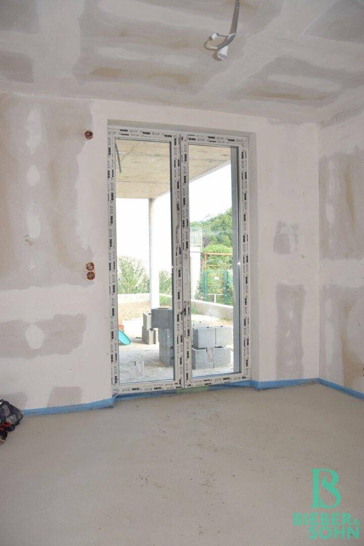 Unfertiger Raum mit großer Glastür, die viel Tageslicht hereinlässt und Zugang nach draußen bietet.