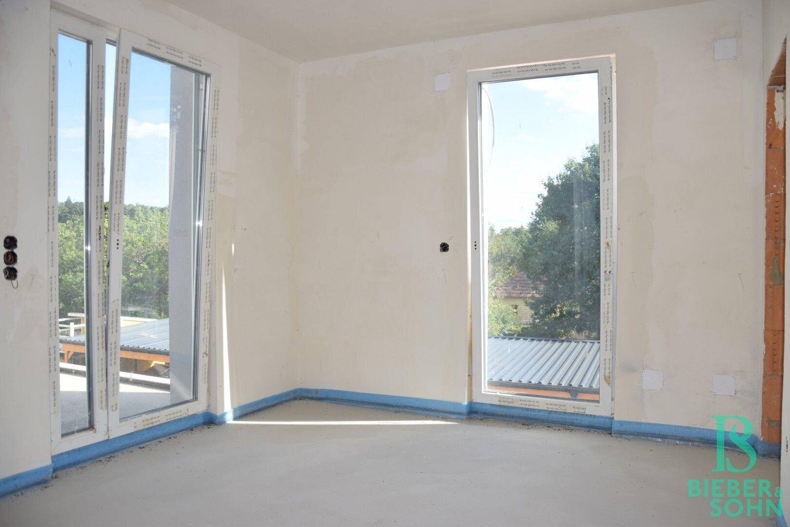 Unfertiger Raum mit großen Fenstern und einer Balkontür, die viel natürliches Licht hereinlassen.
