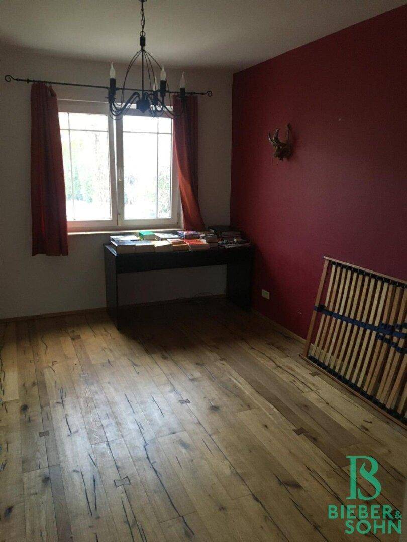 Schlafzimmer mit Holzboden, roter Wand und einem Fenster mit Blick ins Grüne.