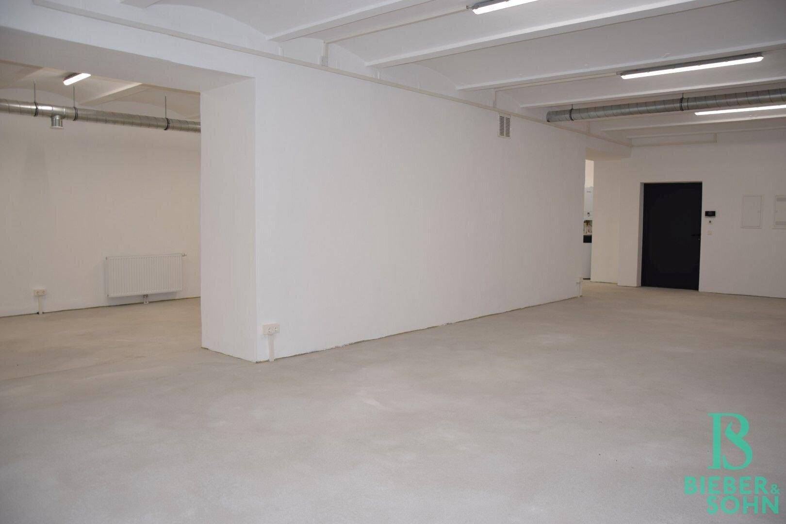 Geräumiger Gewerberaum mit Betonboden, weißen Wänden und einer dunklen Tür im hinteren Bereich.
