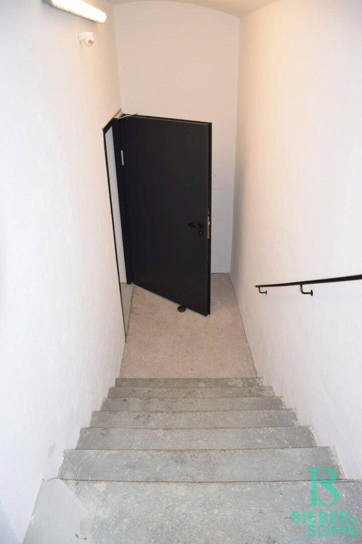 Betontreppe führt nach unten zu einer schwarzen Tür, mit schlichtem schwarzen Handlauf.