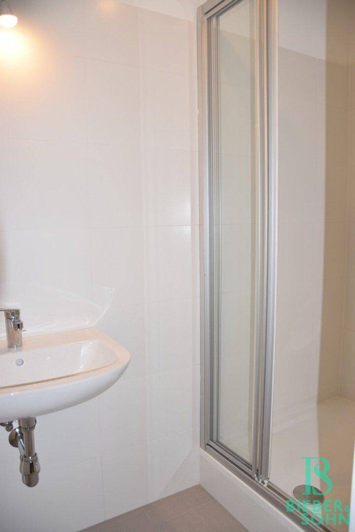 Heller Duschbereich mit Glasabtrennung, kleinem Waschbecken und weißen Wandfliesen.