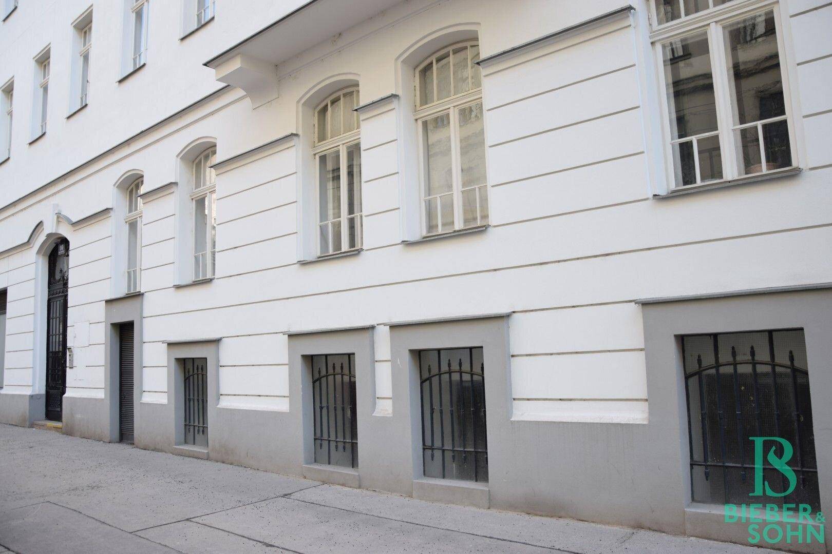 Historische Gebäudefassade mit weißem Putz, grauer Sockelzone und vergitterten Fenstern im Erdgeschoss.