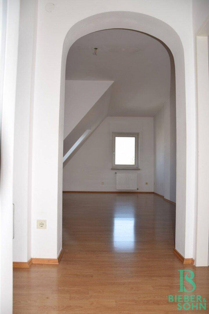 Heller Dachgeschossraum mit Laminatboden, Dachschräge und einem Fenster, verbunden durch einen eleganten Bogen.