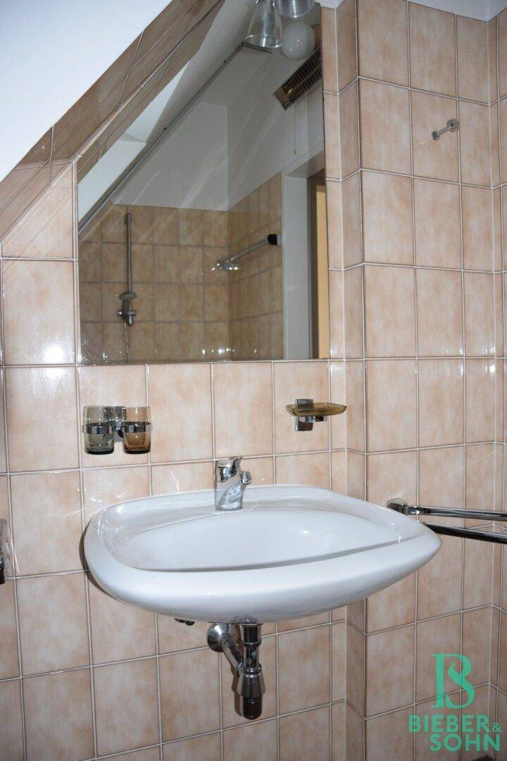 Badezimmer mit Waschbecken, Spiegel und beigefarbenen Fliesen unter einer Dachschräge.