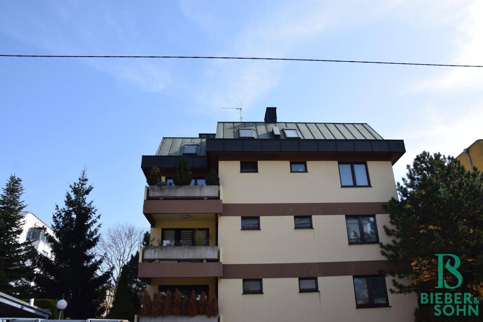 Mehrfamilienhaus mit Balkonen und einem markanten Mansarddach unter blauem Himmel.