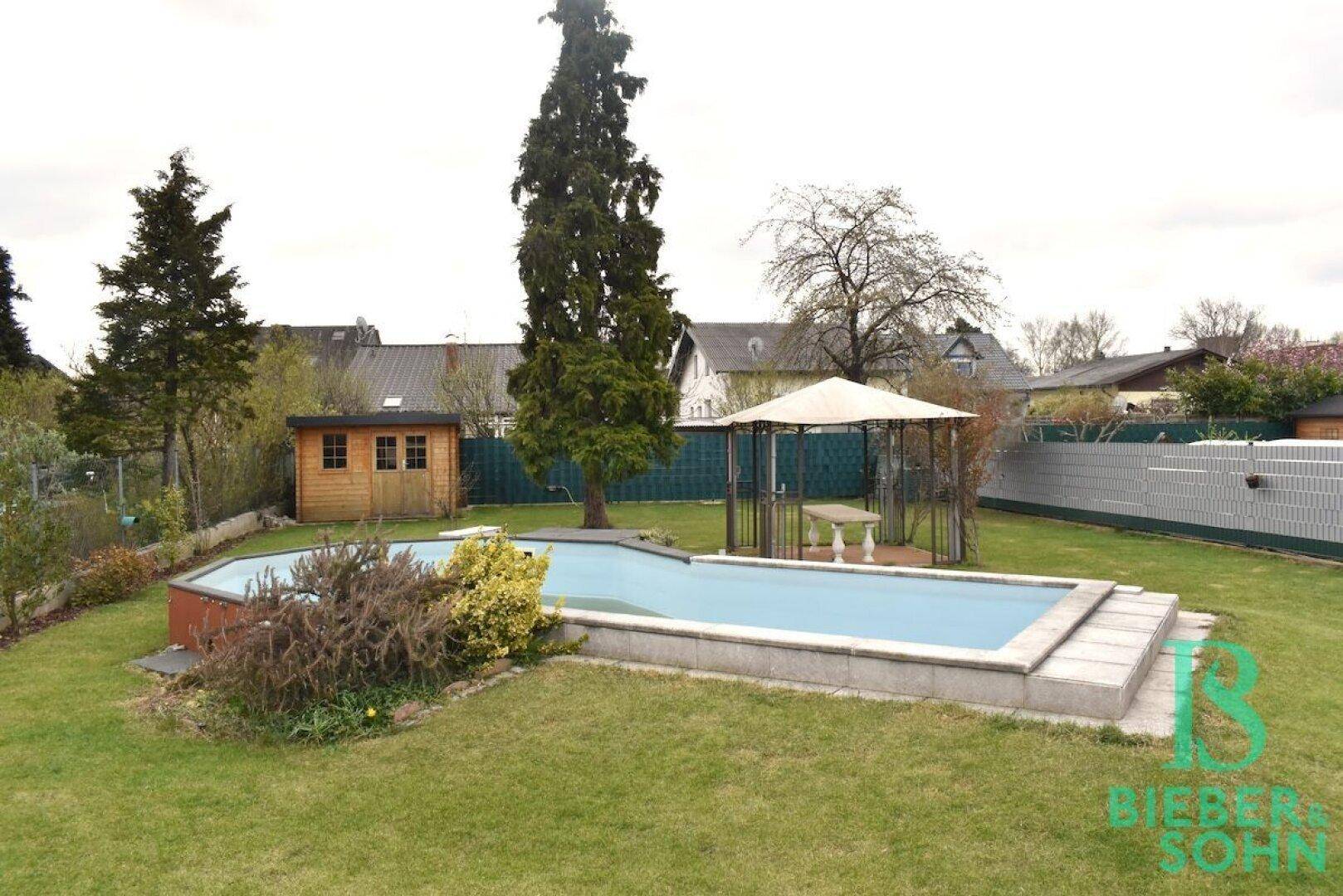 Gartenansicht mit Pool, Gartenlaube, hohen Bäumen und einem kleinen Gartenhaus.