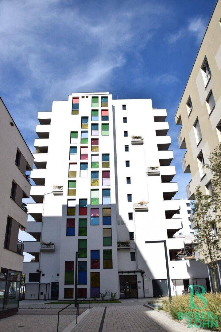 Hohes, modernes Wohngebäude mit weißer Fassade und bunten Fenstern unter blauem Himmel.