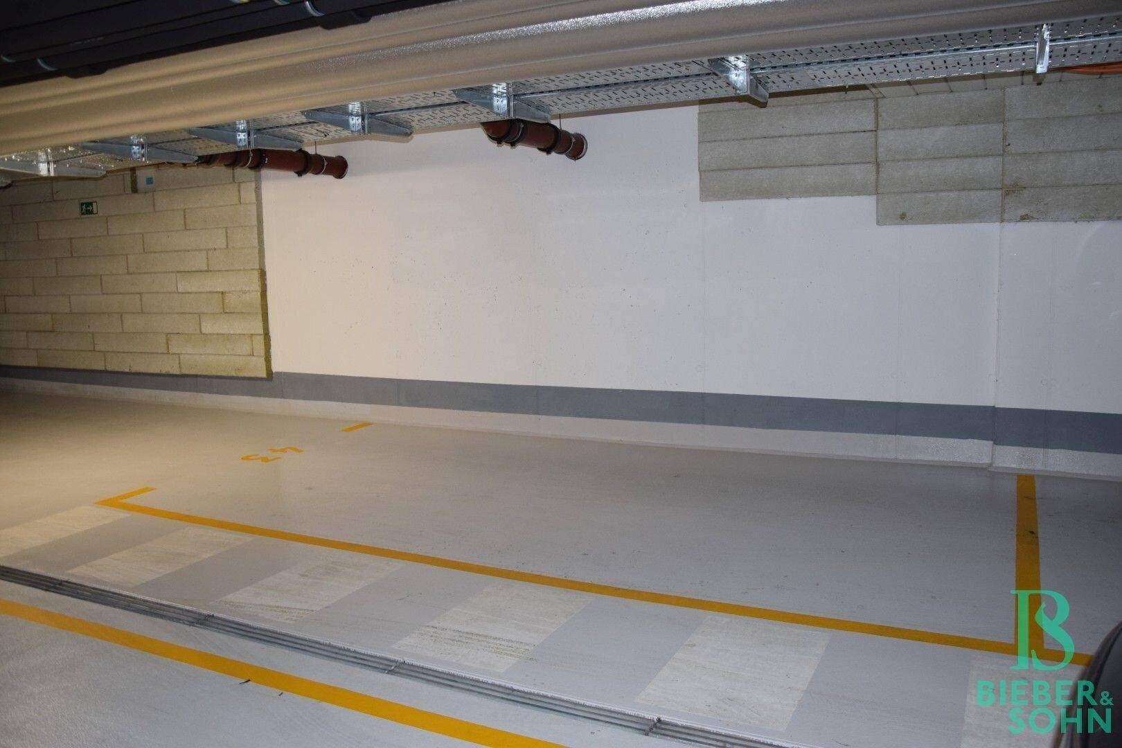 Innenansicht einer Tiefgarage mit markierten Parkplätzen und sichtbaren Installationen an der Decke.