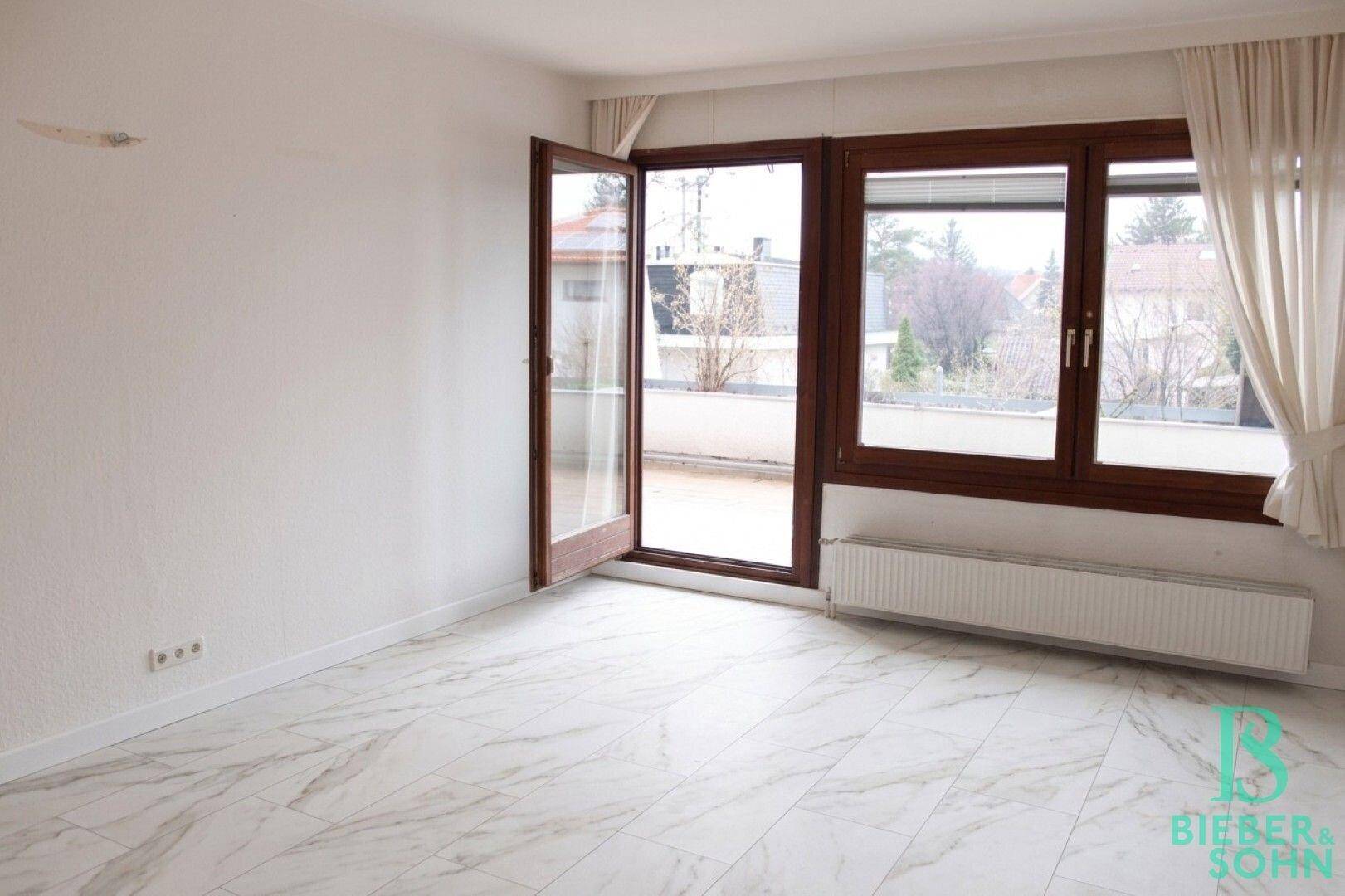 Helles Zimmer mit großen Fenstern, Zugang zum Balkon und Fliesenboden in Marmoroptik.