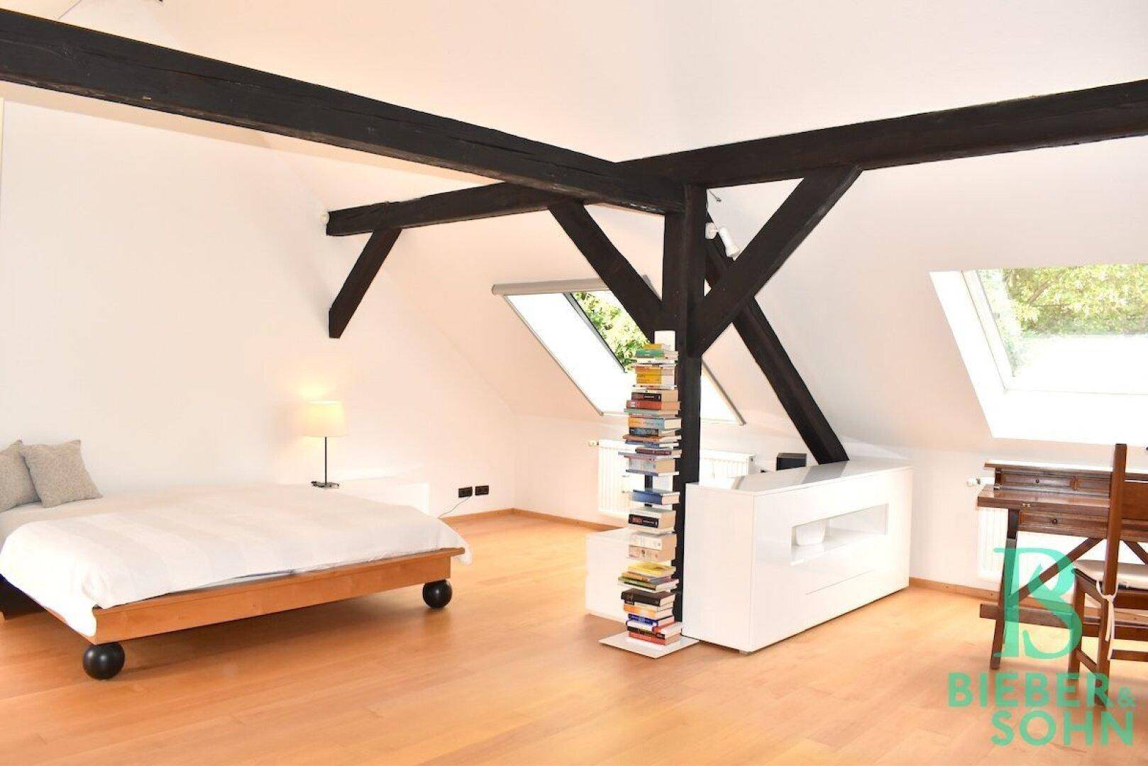 Geräumiges Schlafzimmer im Dachgeschoss mit sichtbaren Holzbalken, Holzboden und Dachfenstern.