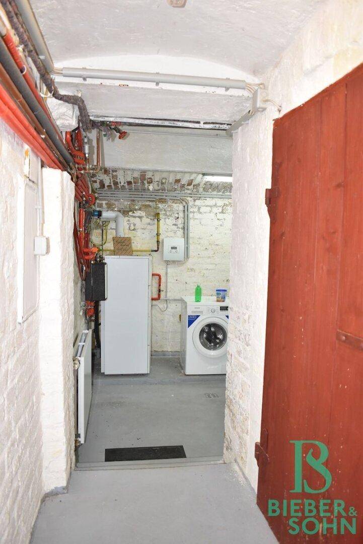 Funktionaler Hauswirtschaftsraum im Keller mit Waschmaschine, Warmwasserbereiter und sichtbaren Leitungen.