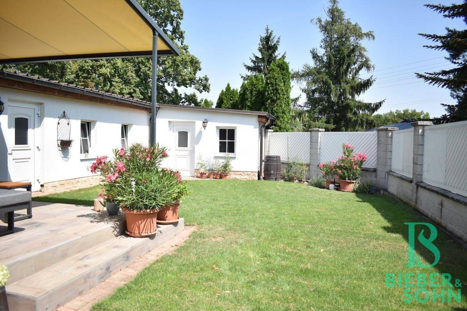 Sonniger Gartenbereich mit Rasen, Terrasse und einem kleinen Nebengebäude im Hintergrund.