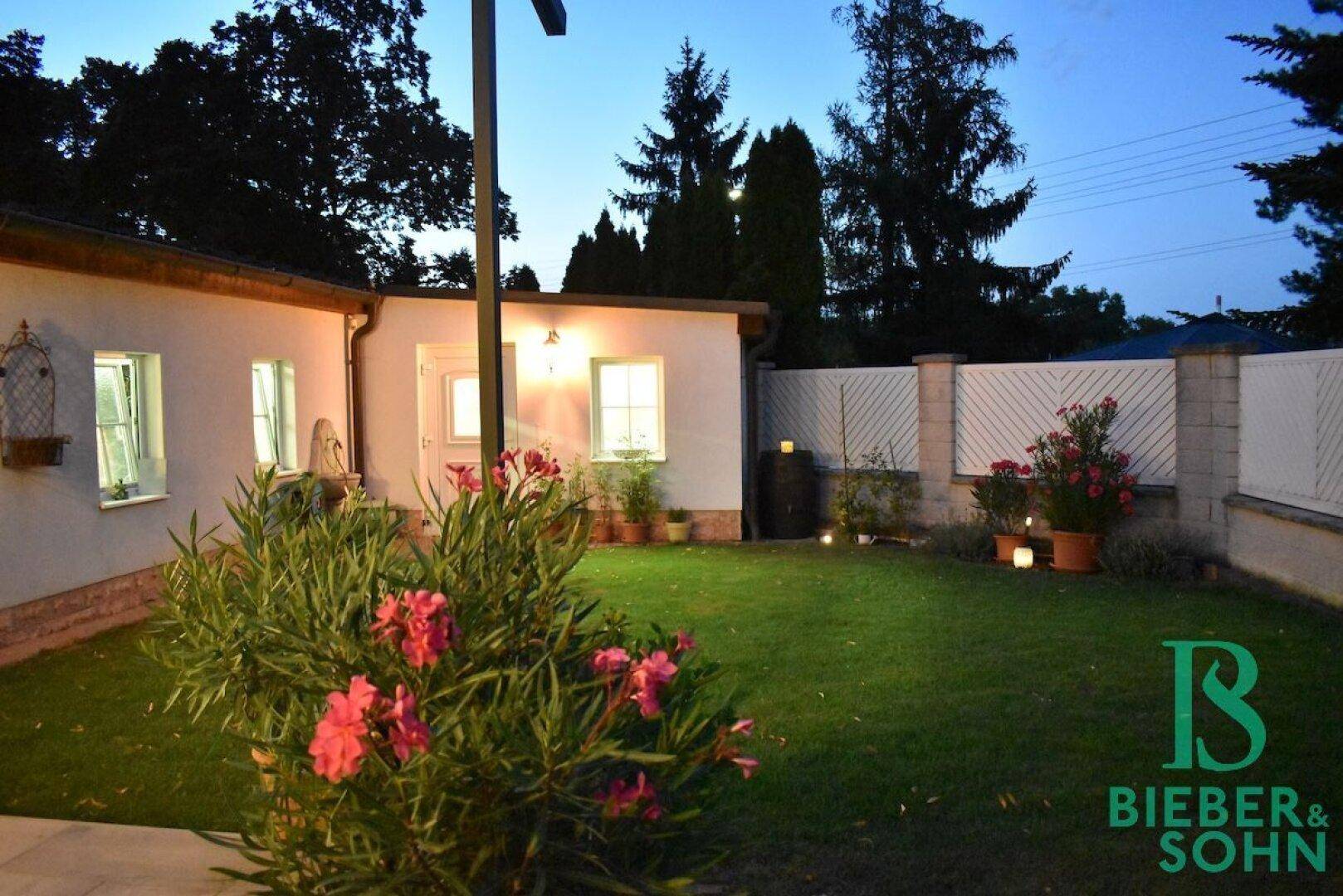 Abendliche Ansicht des Gartens mit blühenden Pflanzen und beleuchteter Hausfassade.