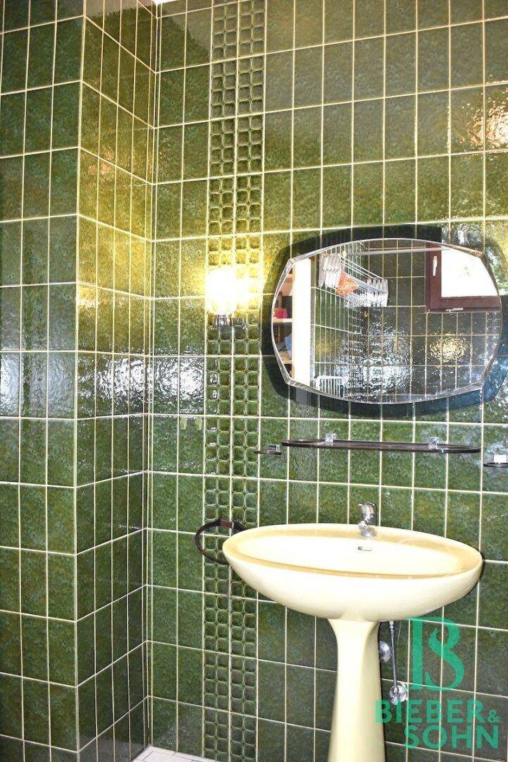Badezimmer mit grünen Fliesen, Standwaschbecken und ovalem Spiegel.