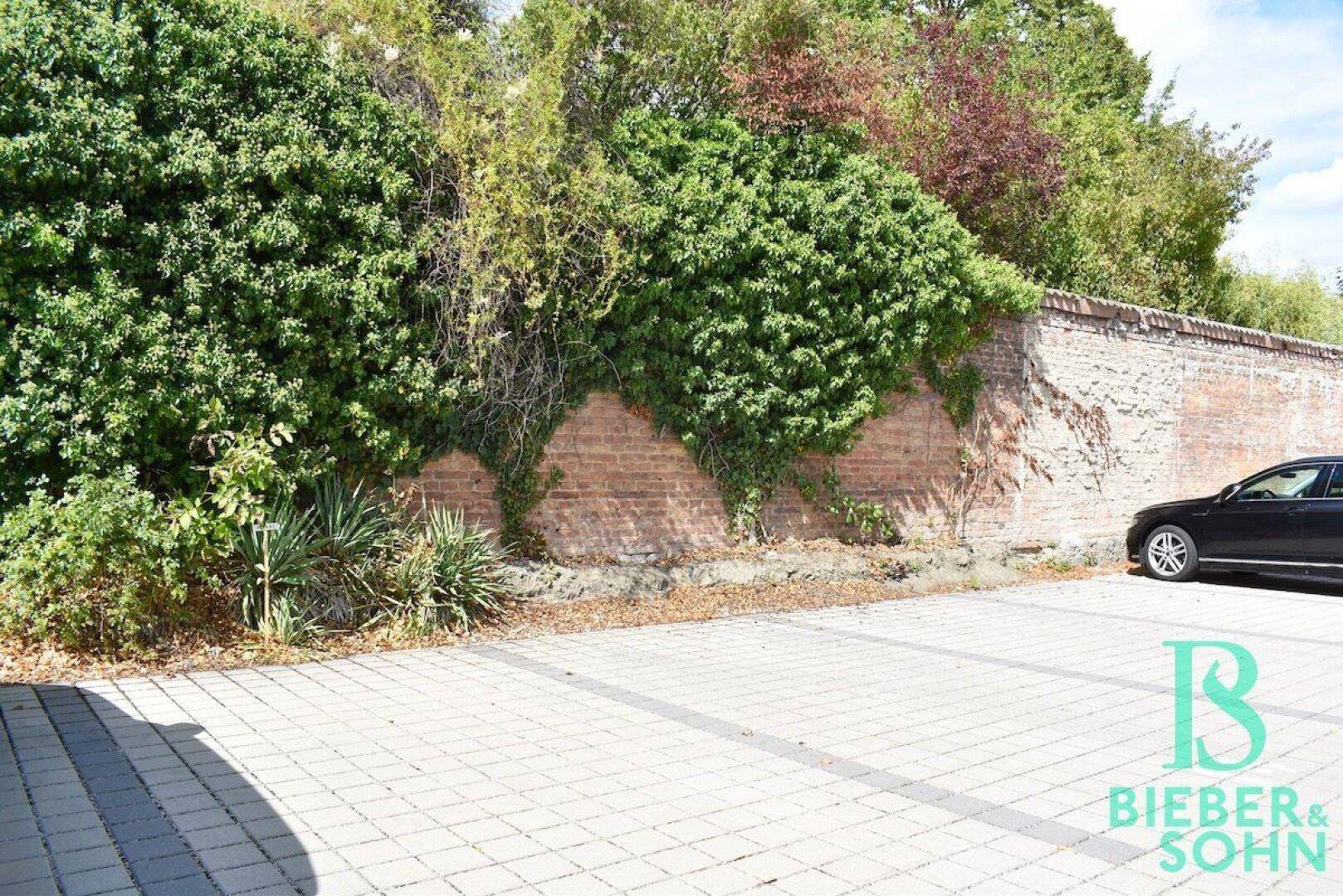 Außenparkplatz mit Pflastersteinen und einer alten Ziegelmauer, umgeben von Grün.