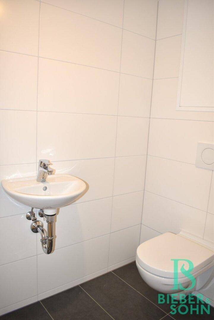 Funktionelles WC mit wandhängender Toilette und kleinem Waschbecken, hell gefliest.