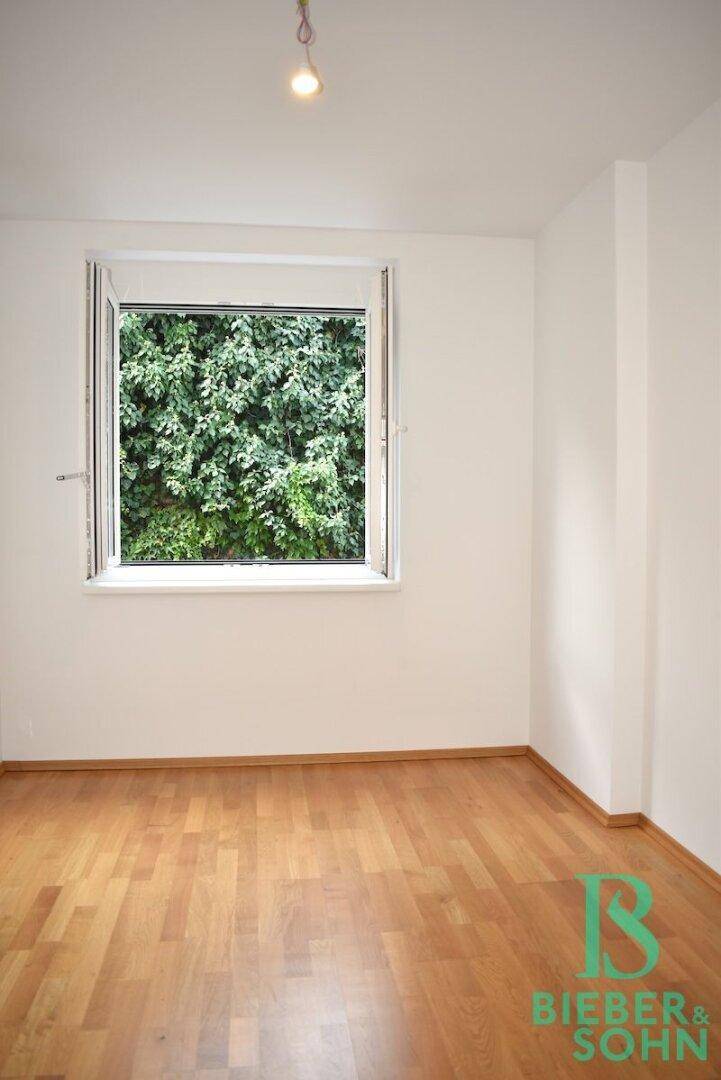 Ruhiges Zimmer mit Holzboden und Fenster zum grünen Außenbereich, ideal als Schlafzimmer.