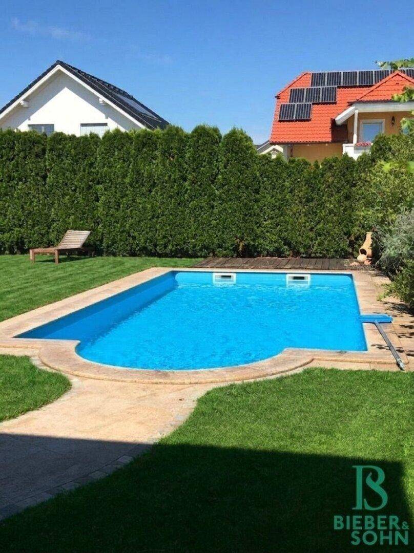 Außenbereich mit einem rechteckigen Swimmingpool, umgeben von Rasen und einer hohen Hecke.