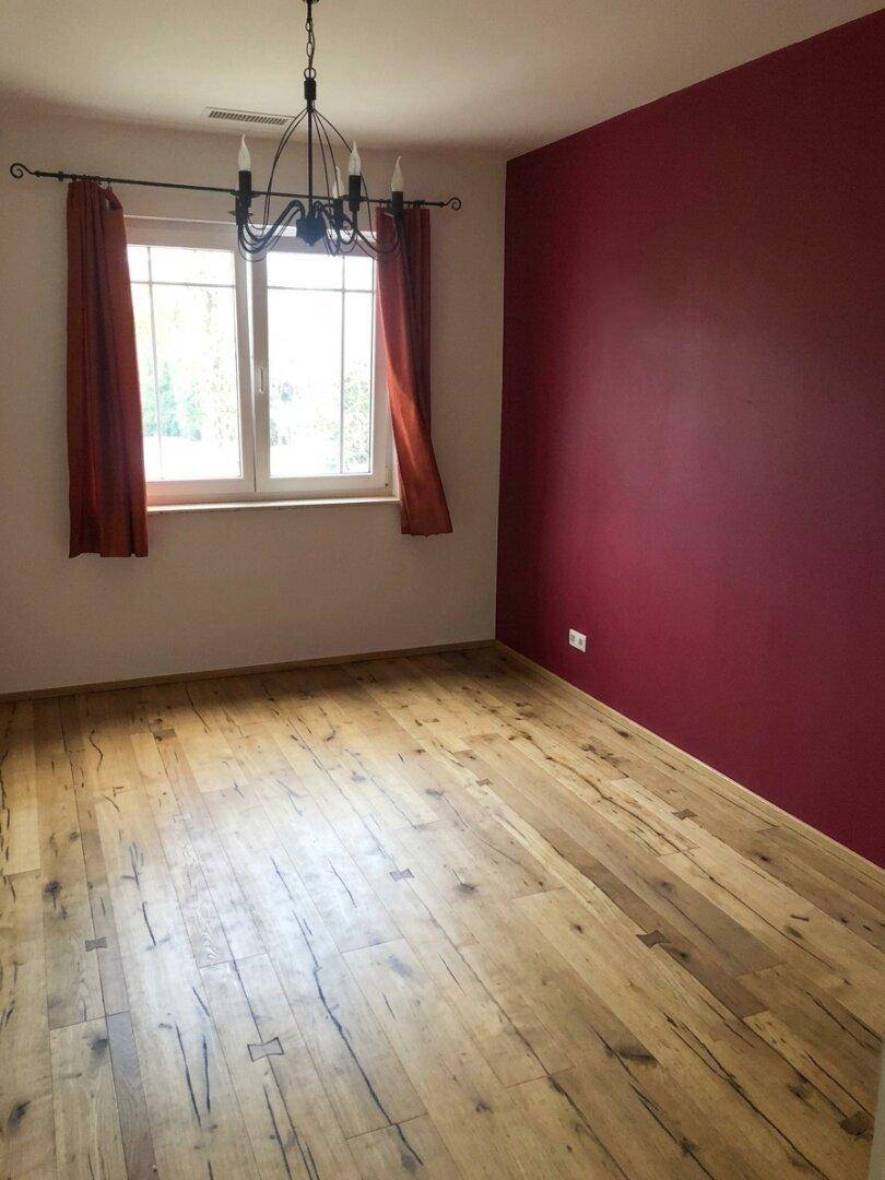 Schlafzimmer mit Holzboden, einer roten Akzentwand und einem Fenster mit Blick ins Grüne.