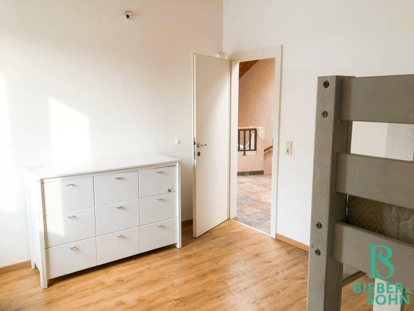 Schlafzimmer mit Holzboden, weißer Kommode und Zugang zu einem weiteren Raum.