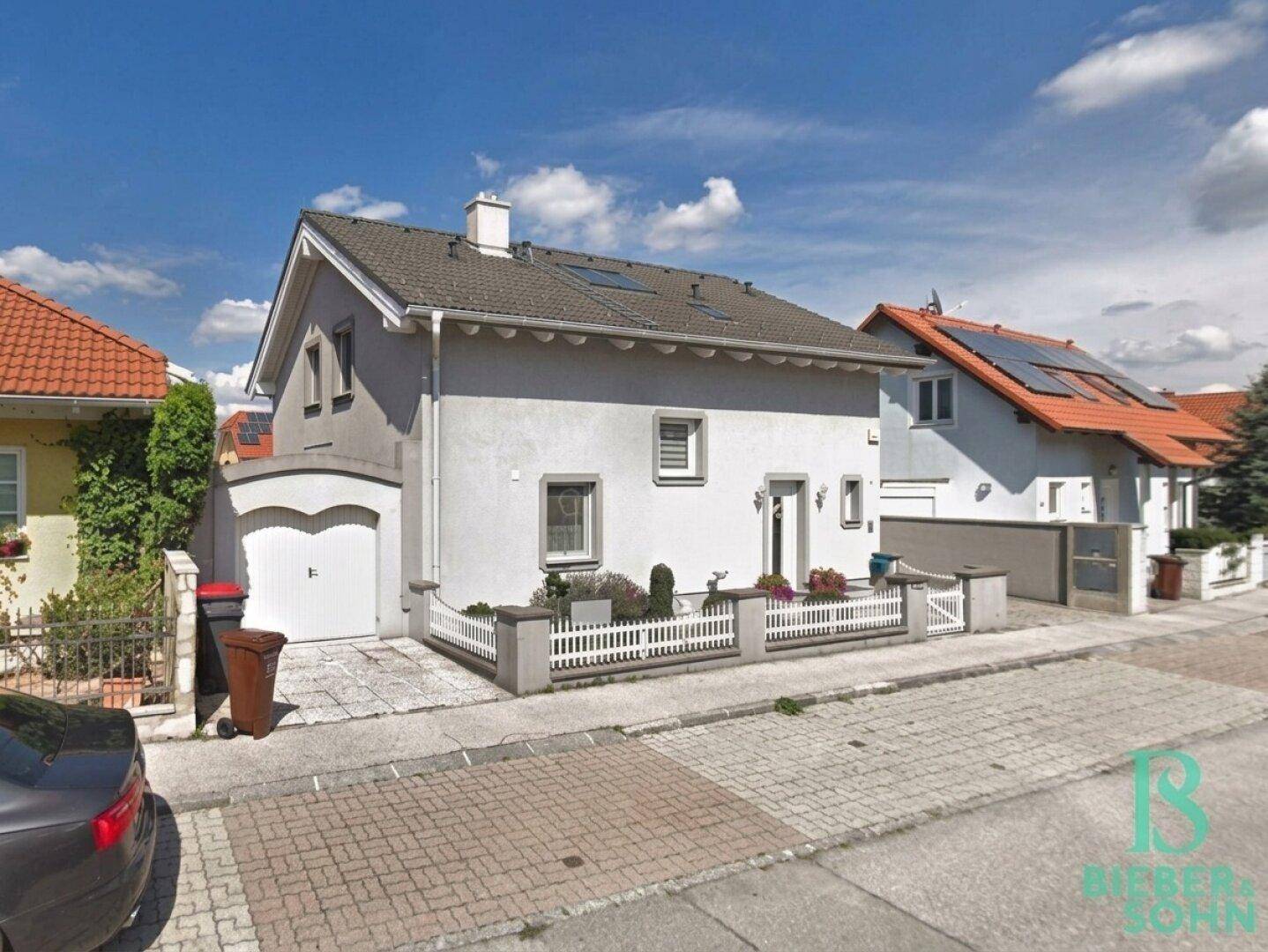 Einfamilienhaus mit grauer Fassade, weißem Zaun und angrenzender Garage an der Straße.