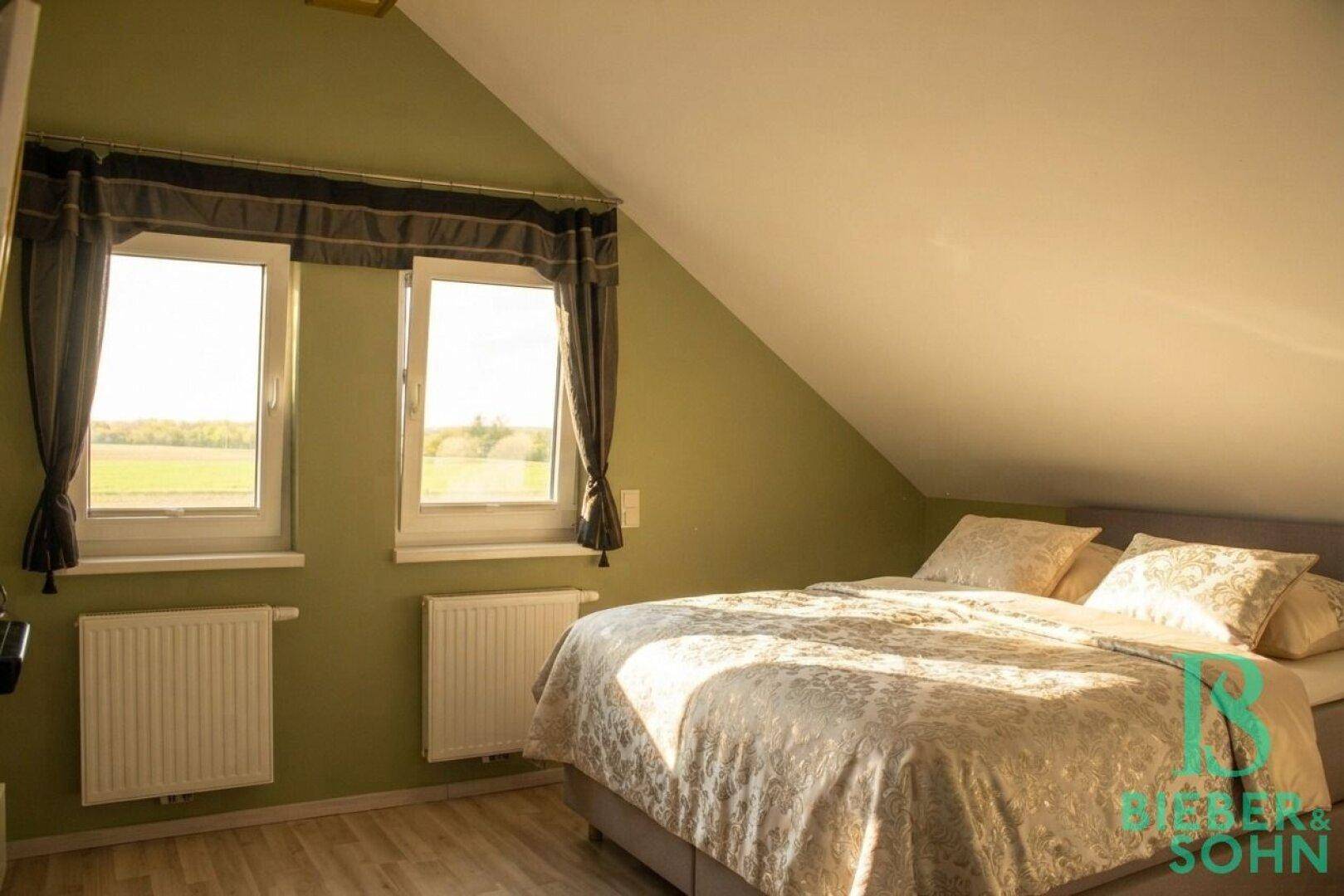 Helles Schlafzimmer im Dachgeschoss mit grünen Wänden, Holzboden und weitem Blick ins Grüne.