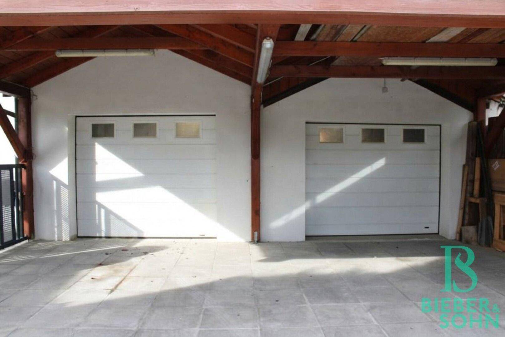 Zwei Garagentore unter einem robusten Holzcarport mit gepflastertem Vorplatz.