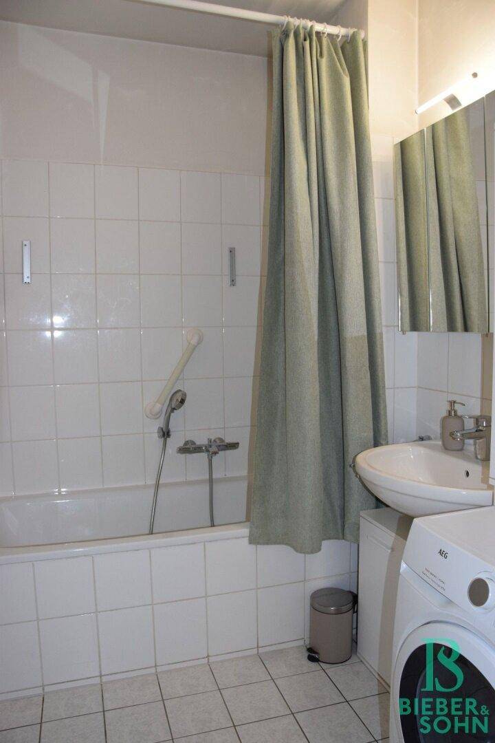 Funktionales Badezimmer mit weißen Fliesen, Badewanne mit Duschvorhang und Waschmaschinenanschluss.