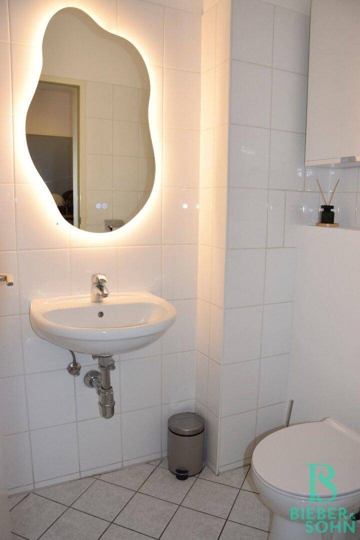 Modernes Gäste-WC mit beleuchtetem Spiegel, Waschbecken und weißen Fliesen.