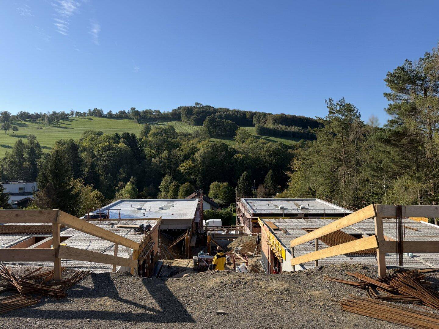 Baustelle mit im Bau befindlichen Gebäuden und weitem Blick auf die idyllische grüne Hügellandschaft unter blauem Himmel.