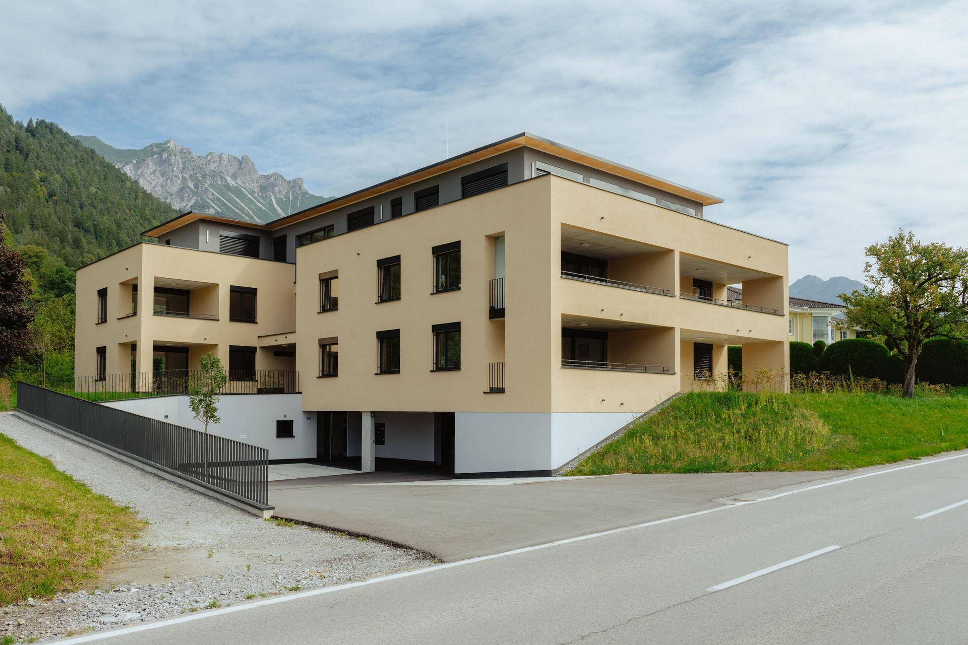 Moderne Wohnanlage mit gelber Fassade, Balkonen und Tiefgaragenzufahrt in bergiger Umgebung.