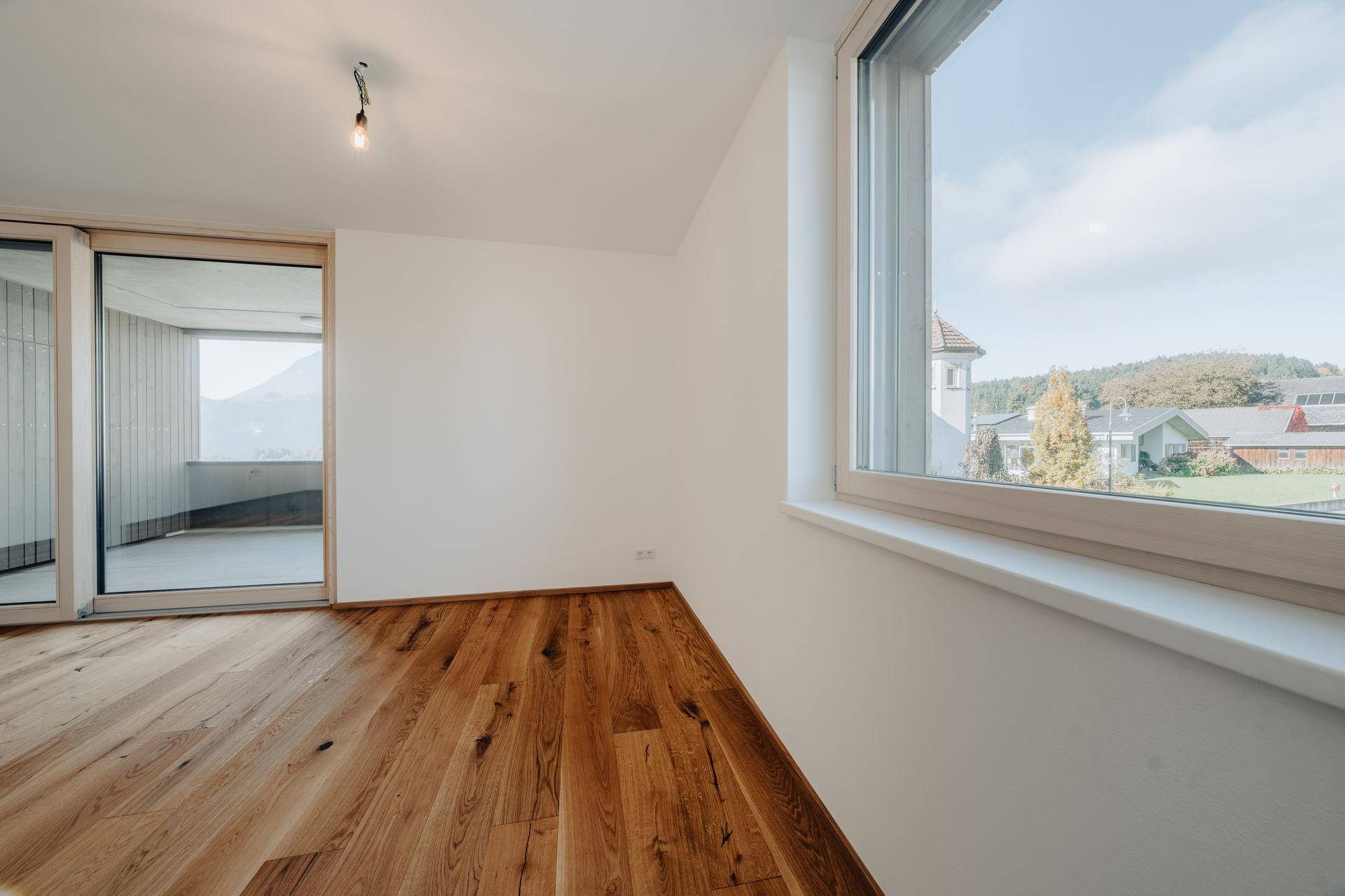 Heller Raum mit Holzboden, großem Fenster und Zugang zu einem Außenbereich mit Bergblick.