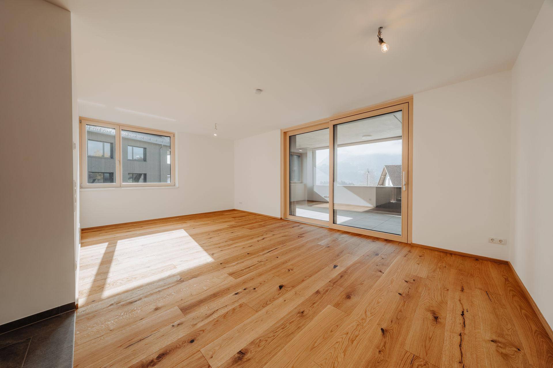 Heller Wohnraum mit breiten Holzplankenböden, großen Fenstern und Zugang zu einem Außenbereich.