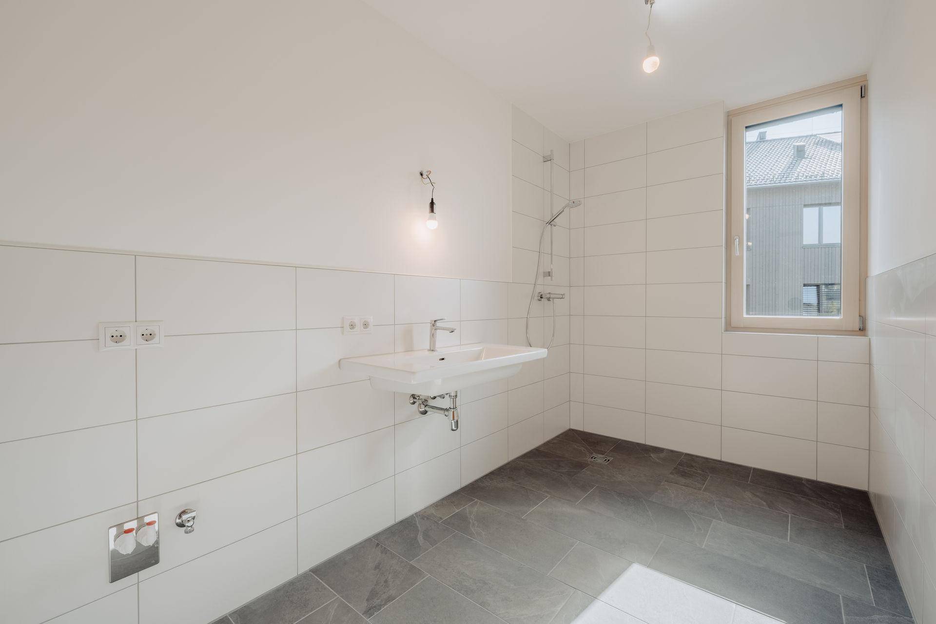 Modernes Badezimmer mit weißen Wandfliesen, grauem Boden und einem Fenster für Tageslicht.