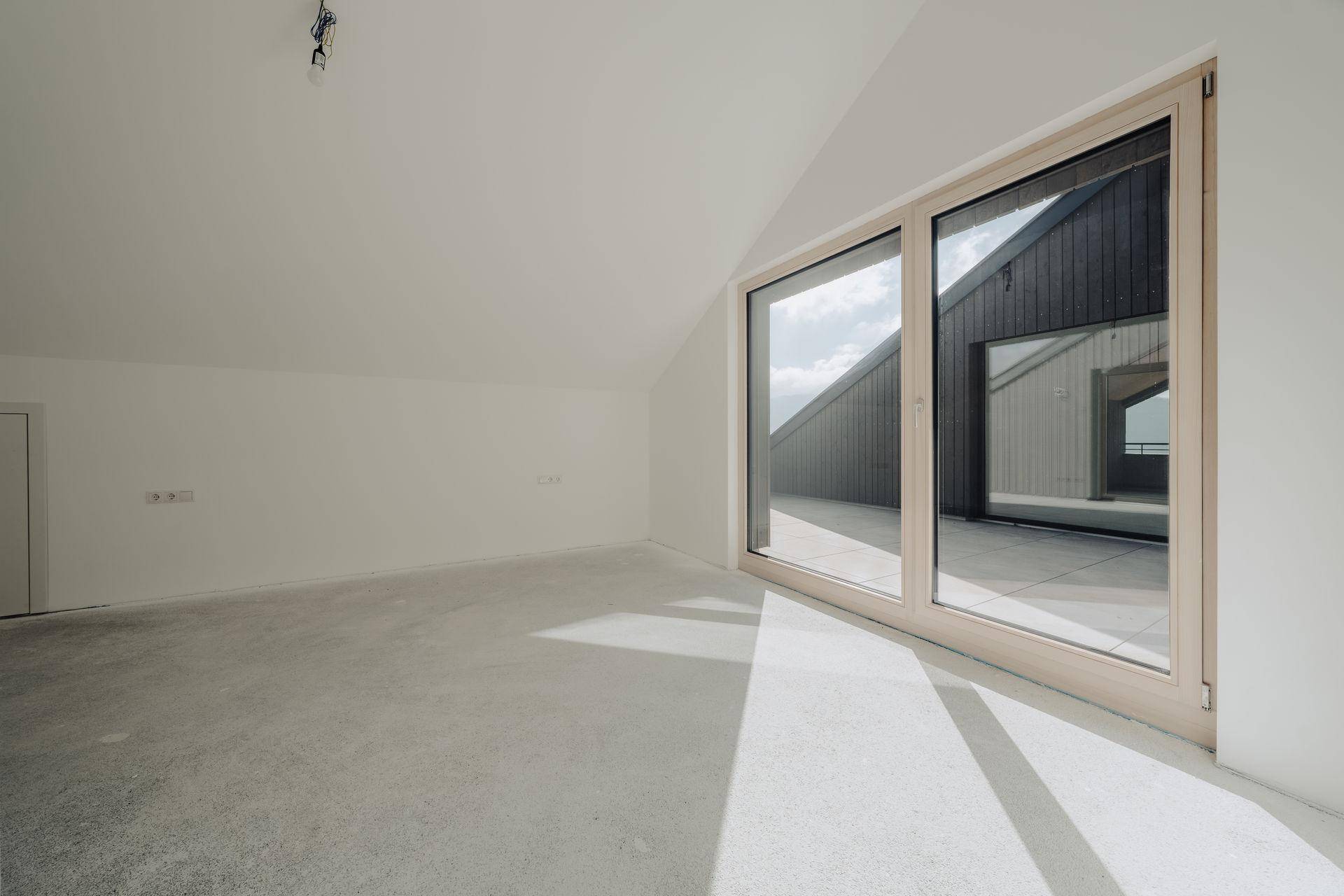 Lichtdurchfluteter, unmöblierter Raum mit großen Fenstern und schrägen Decken im Neubau.