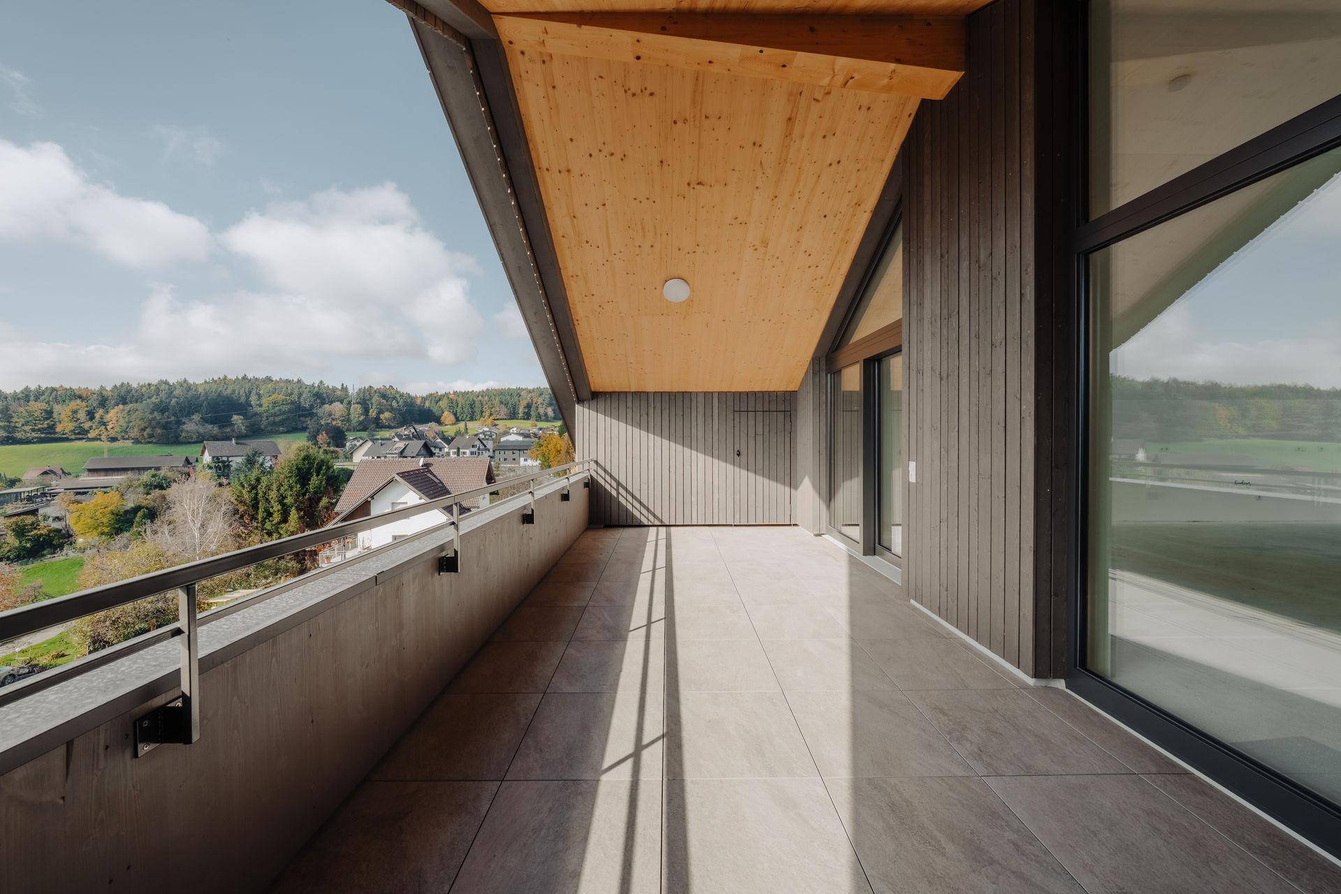 Langer Balkon mit Fliesenboden und Holzdach, bietet weite Aussicht auf die grüne Umgebung.