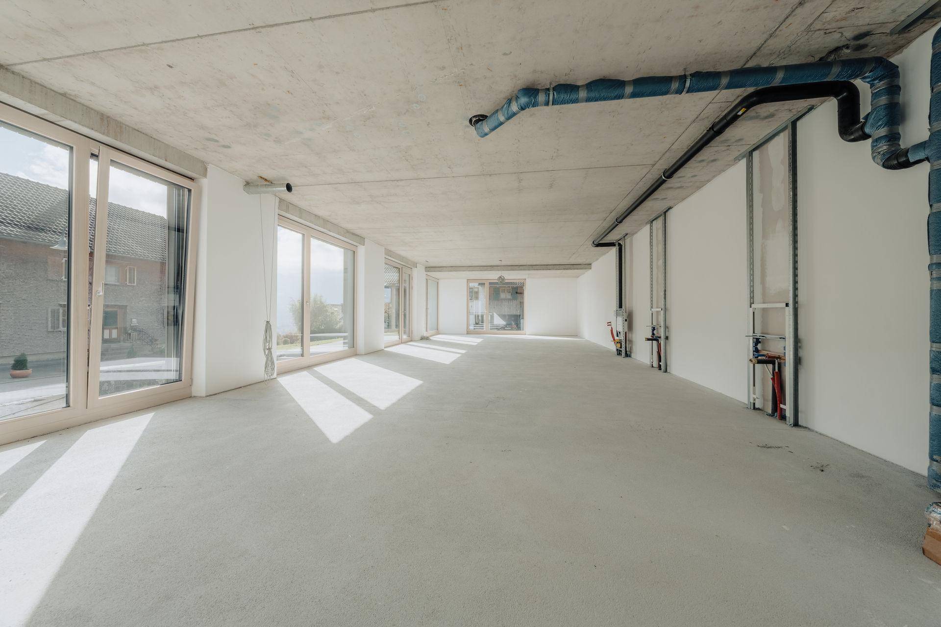 Großer, unfertiger Innenraum mit Betonwänden und -decke, großen Fenstern und sichtbaren Installationen.