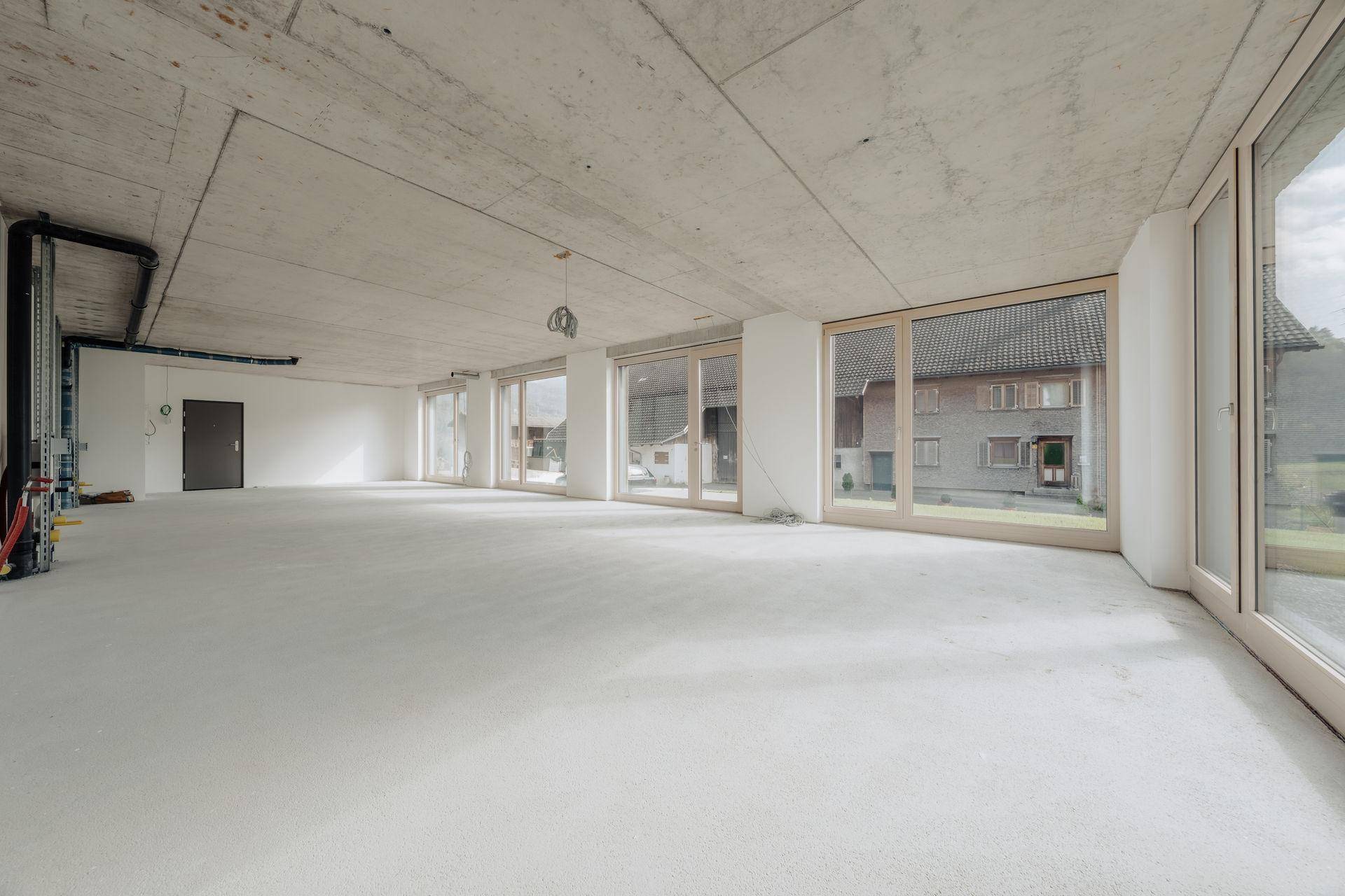 Unfertiger Gewerberaum mit Betonoberflächen, großen Fensterfronten und sichtbaren Installationen.
