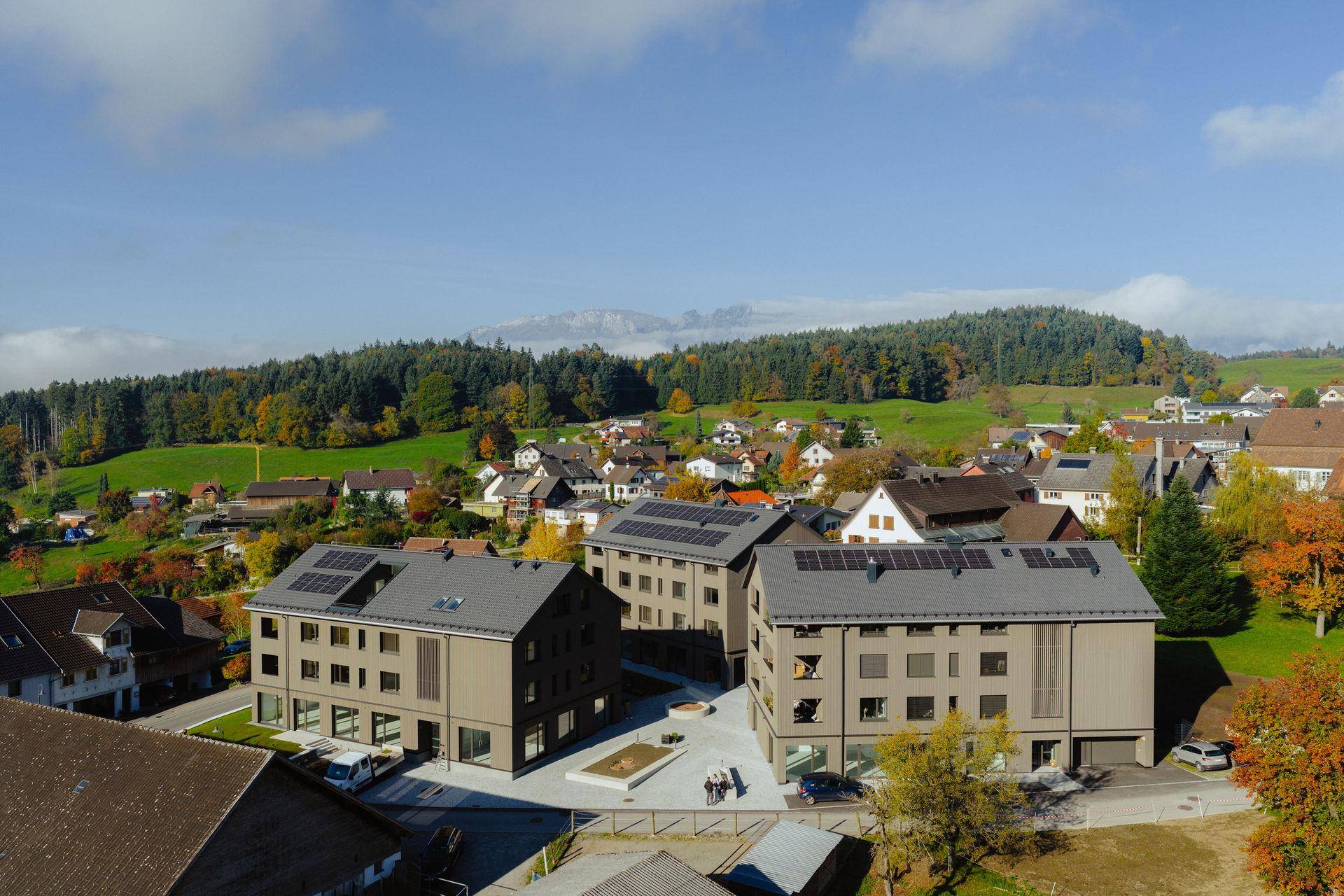Luftaufnahme von modernen Neubauten in einer ländlichen Umgebung mit bewaldeten Hügeln und Bergen im Hintergrund.