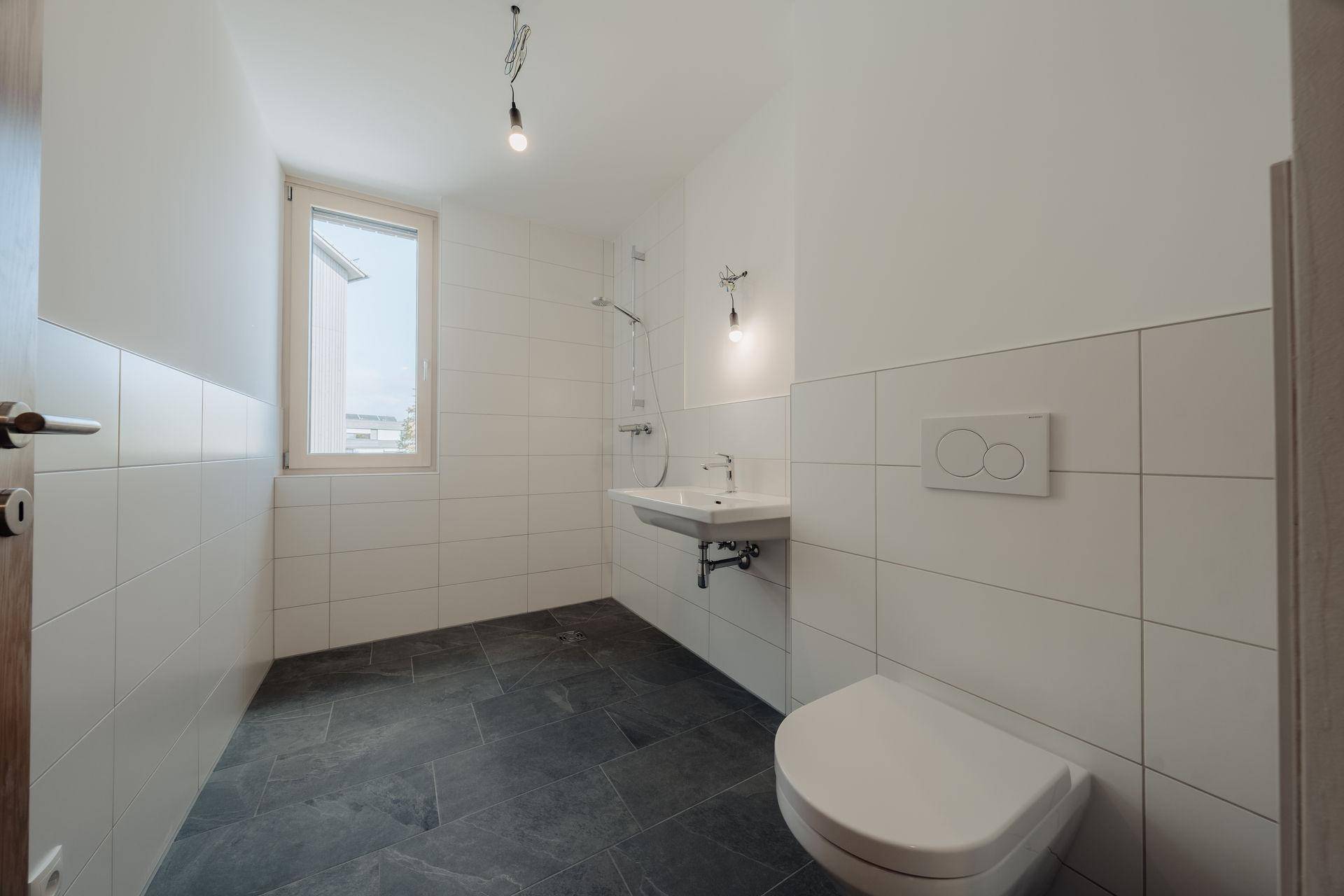 Modernes Badezimmer mit weißen Wandfliesen, dunklem Boden und wandhängendem WC, ausgestattet mit Fenster und Dusche.