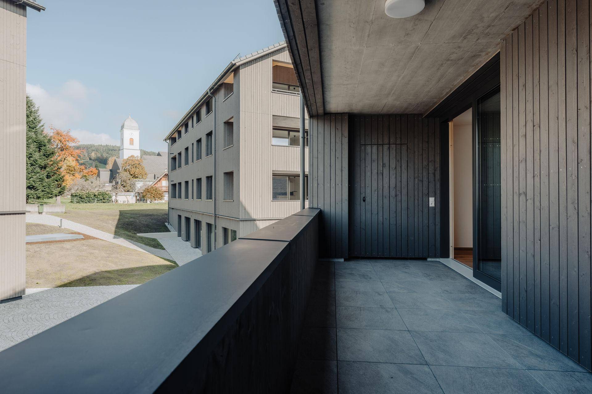 Moderner Balkon mit grauen Fliesen und dunklem Geländer, bietet einen Blick auf die Umgebung und einen Kirchturm.